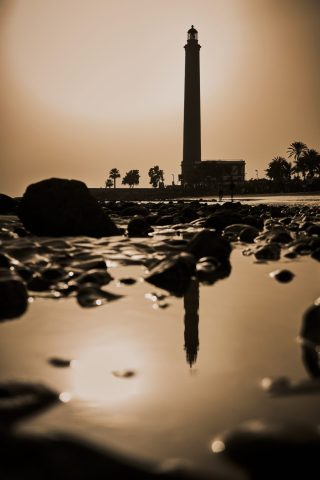 Silhouette d'un grand phare côtier et son reflet dans l'eau d'une plage rocheuse au crépuscule