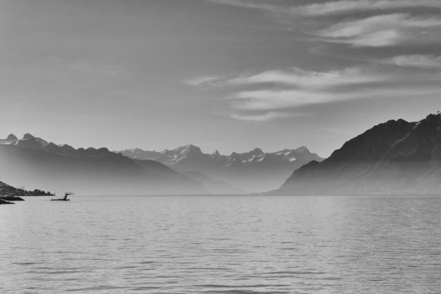 Photographie en noir et blanc d'un lac paisible, de montagnes lointaines et d'un plongeoir isolé