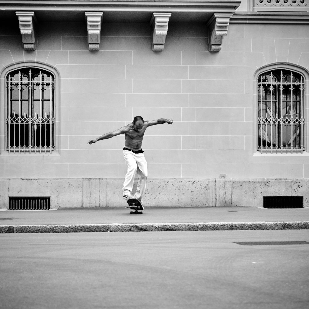 Homme torse nu faisant du skateboard sur un trottoir devant un bâtiment classique aux fenêtres cintrées, photographie en noir et blanc.