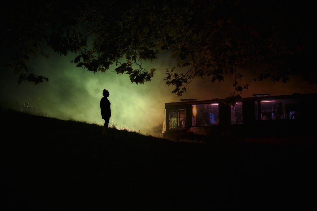 Silhouette d'une personne de nuit observant un ancien bus illuminé au cœur d'un brouillard coloré.