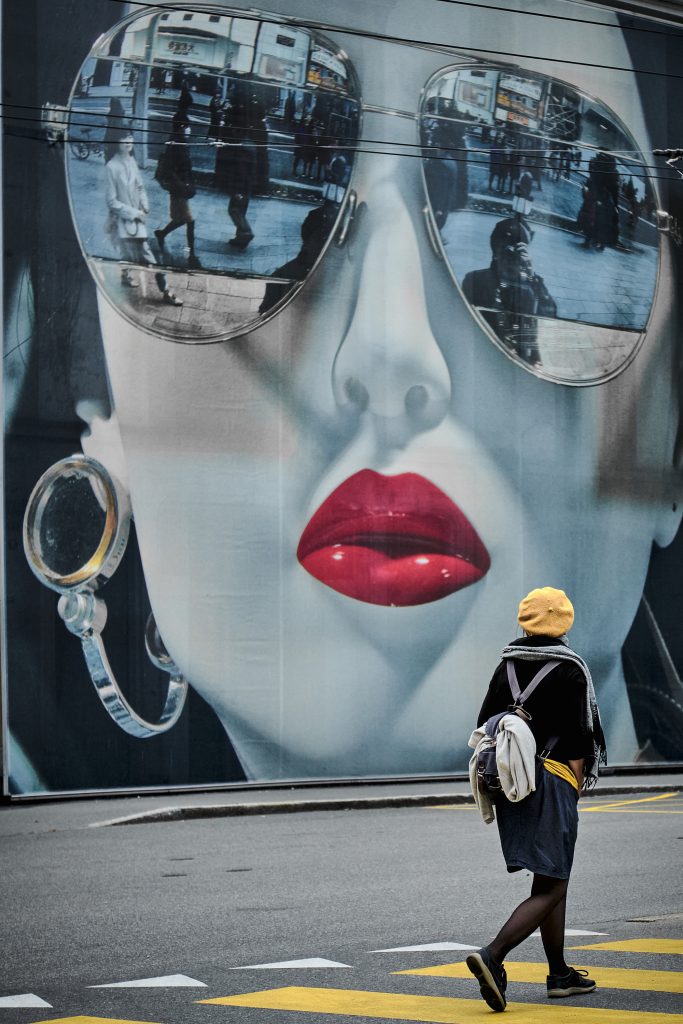 Photographie de rue d'une personne en béret jaune passant devant une fresque murale géante d'un visage avec des lunettes de soleil.
