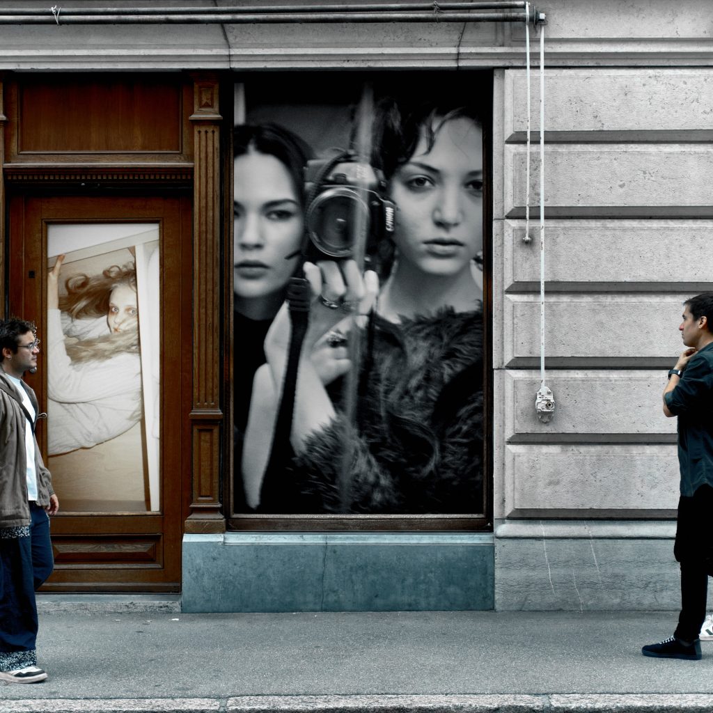 Deux passants sur un trottoir s'observant devant la vitrine d'un bâtiment décorée de grands portraits photographiques en noir et blanc.