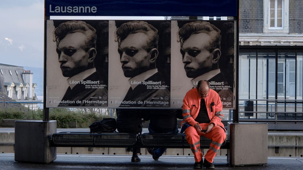 Une personne en tenue de travail orange est assise sur un banc à la gare de Lausanne, regardant son téléphone devant une affiche d'exposition artistique.