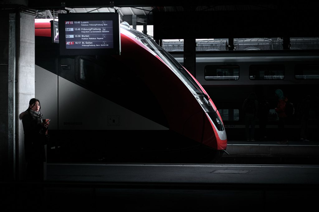 Photographie urbaine contrastée d'une femme attendant dans la pénombre sur le quai d'une gare en Suisse, à côté d'un train moderne rouge et blanc sous un panneau d'affichage des départs.
