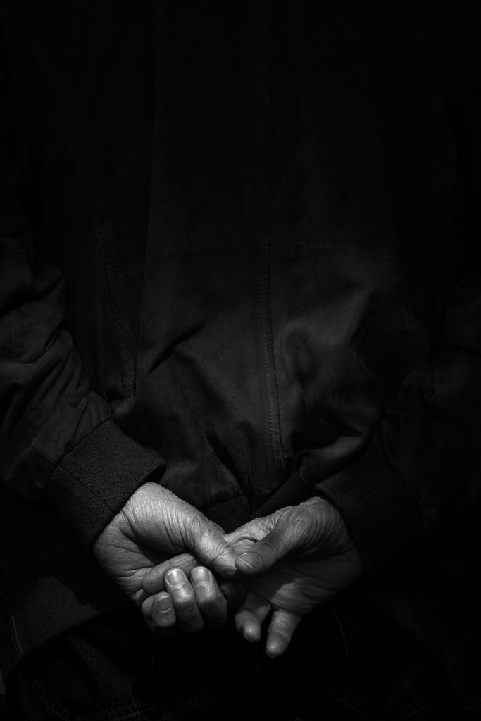 Photographie de rue en noir et blanc et en clair-obscur, offrant un gros plan détaillé sur les mains croisées dans le dos d'une personne portant une veste sombre.