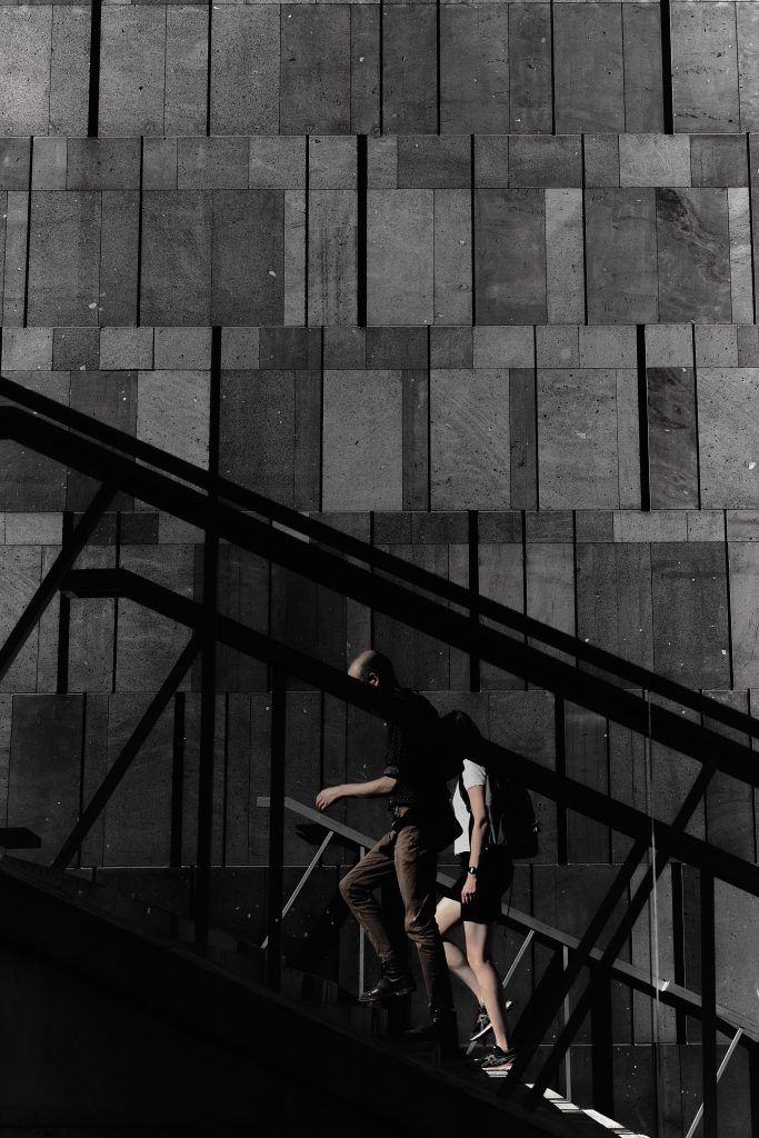 Photographie urbaine de deux personnes en silhouette montant des escaliers en métal devant un mur géométrique en pierre texturée.