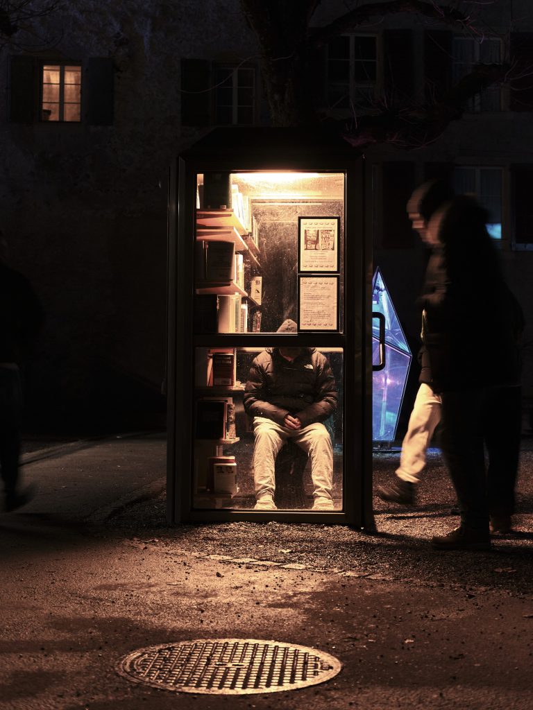 Scène de nuit d'une personne assise dans une cabine téléphonique publique illuminée et transformée en boîte à livres ou bibliothèque de rue.