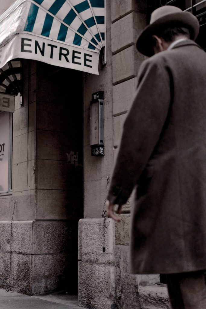 Photographie de rue d'une personne en manteau long et chapeau marchant devant un bâtiment avec un auvent rayé portant l'inscription "ENTREE", ambiance vintage.