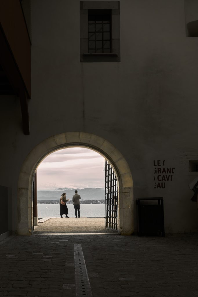Vue encadrée par une grande arche en pierre d'un couple admirant un vaste lac et des montagnes au loin, près d'un mur portant l'inscription "LE GRAND CAVEAU".