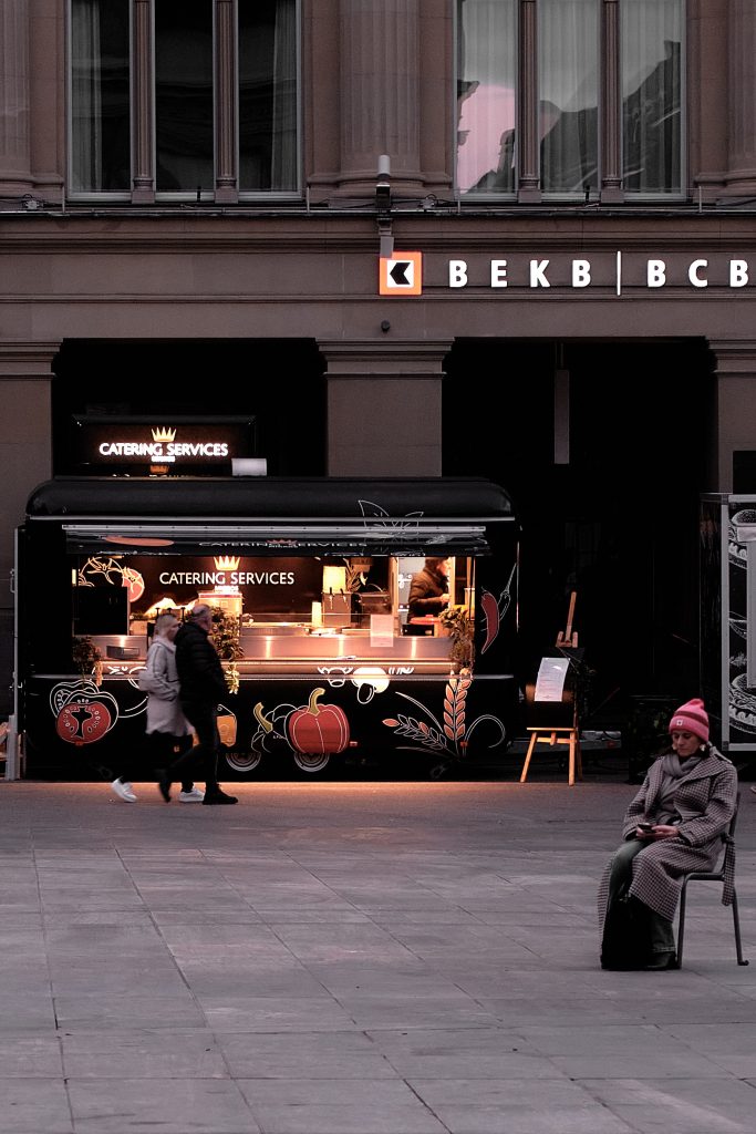 Scène de rue nocturne avec un food truck, des passants et une femme assise regardant son téléphone."