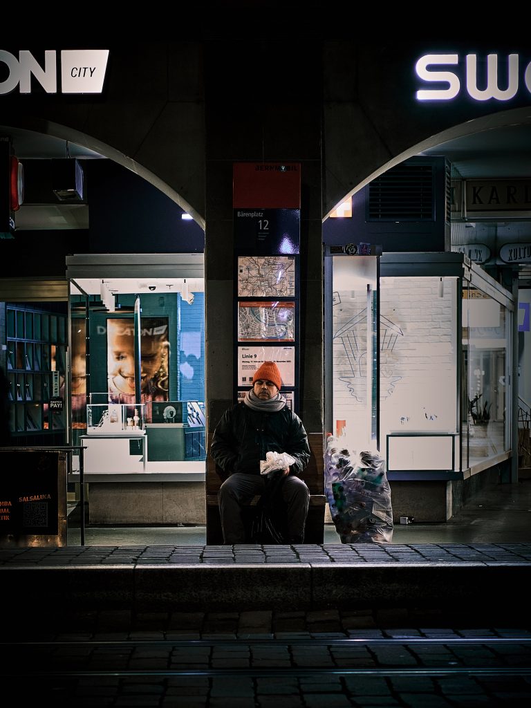 Homme assis sur un banc la nuit entre deux vitrines de magasins éclairées, portant un bonnet orange et ayant à ses côtés un grand sac en plastique rempli de bouteilles vides.