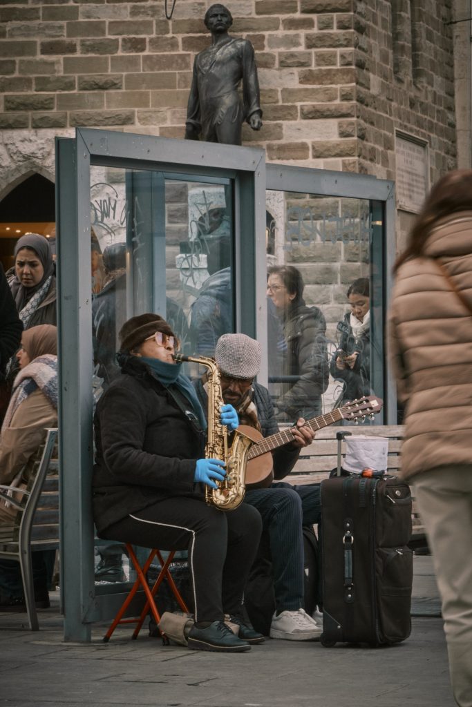 Deux musiciens de rue jouant du saxophone et de la guitare classique, assis devant un abribus vitré où attendent des passants en manteaux d'hiver, sous une statue en bronze.