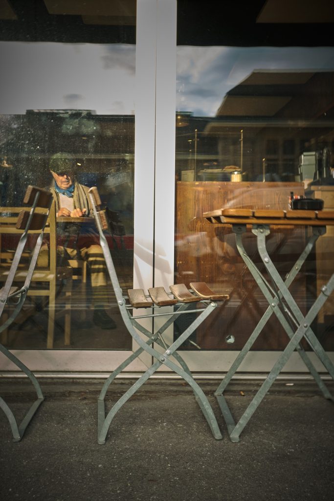 Vue à travers la vitre d'un café montrant un homme âgé avec une casquette assis à l'intérieur, avec des tables et chaises de terrasse pliantes vides au premier plan à l'extérieur.
