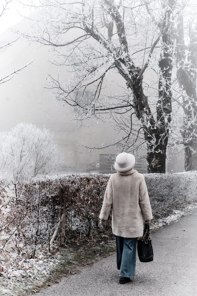 Personne vue de dos portant un manteau duveteux clair et un chapeau assorti, marchant sur un chemin bordé d'arbres et d'une haie entièrement recouverts de givre blanc.