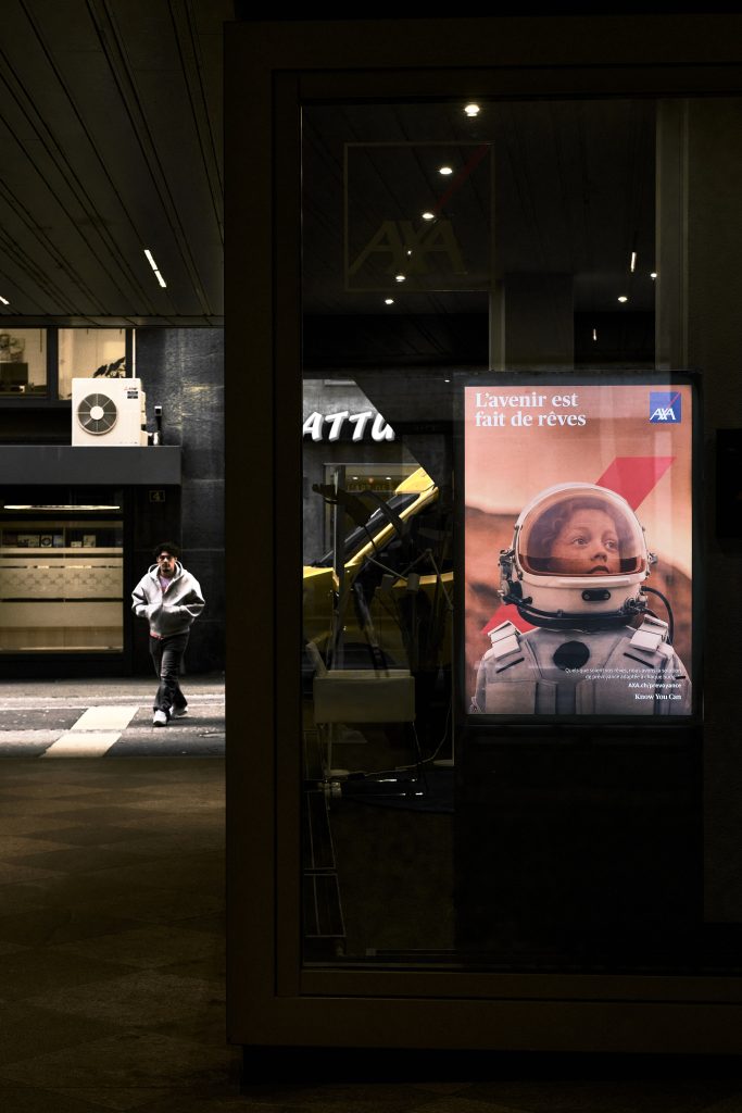 Panneau publicitaire lumineux dans la rue la nuit affichant un enfant en tenue d'astronaute, contrastant avec la silhouette d'un passant marchant dans une ruelle sombre en arrière-plan.