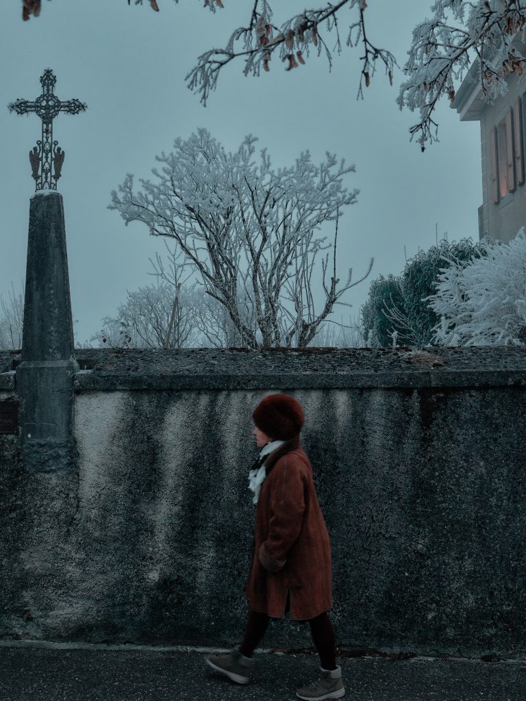 Personne portant un manteau rouille et un bonnet d'hiver marchant le long d'un vieux mur en pierre surmonté d'une croix métallique monumentale, avec des arbres givrés sous un ciel gris.