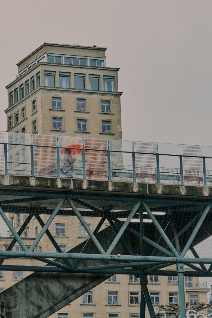 Personne tenant un parapluie orange marchant sur une passerelle piétonne métallique avec un grand bâtiment urbain en arrière-plan sous un ciel gris.