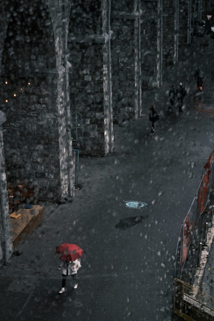 Vue en plongée à travers une vitre couverte de gouttes de pluie montrant une personne avec un parapluie rouge marchant dans une ruelle sombre bordée d'arches en pierre.