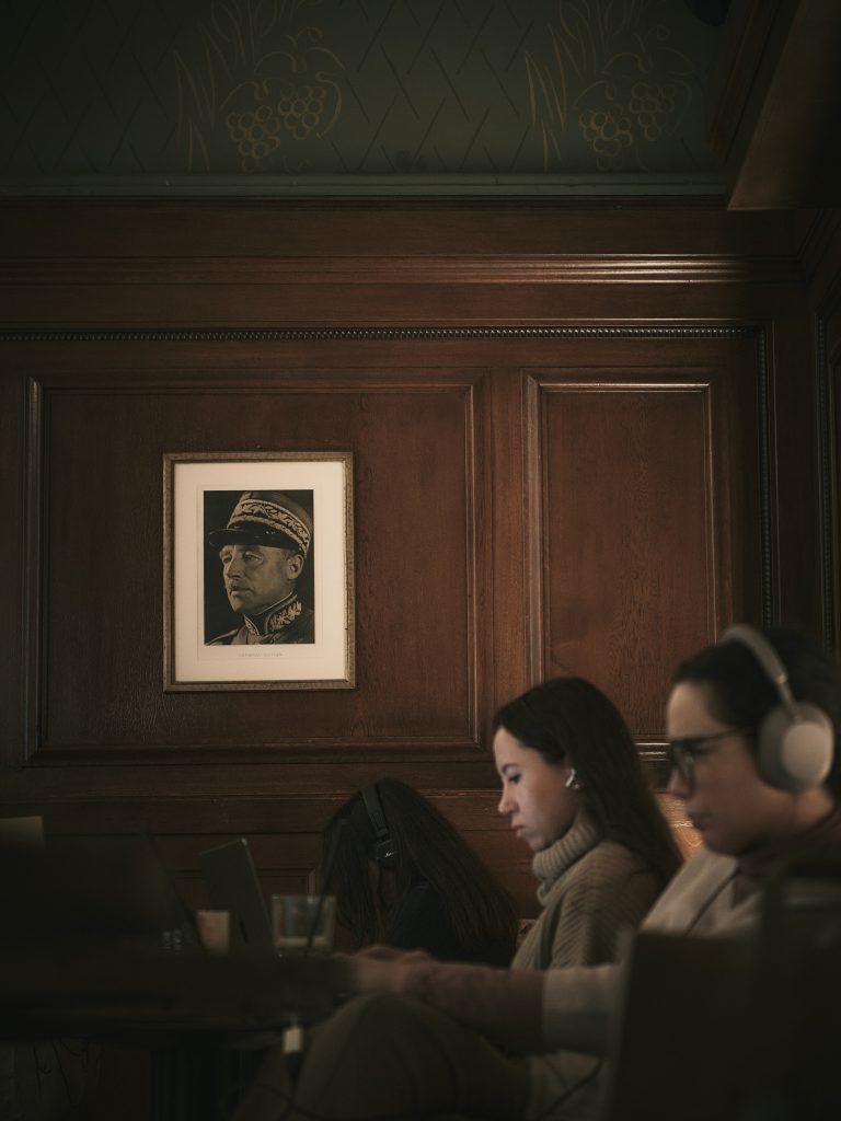 Deux personnes portant des casques audio travaillant sur des ordinateurs portables dans une pièce chaleureuse aux murs en boiseries sombres, sous un portrait militaire historique encadré.