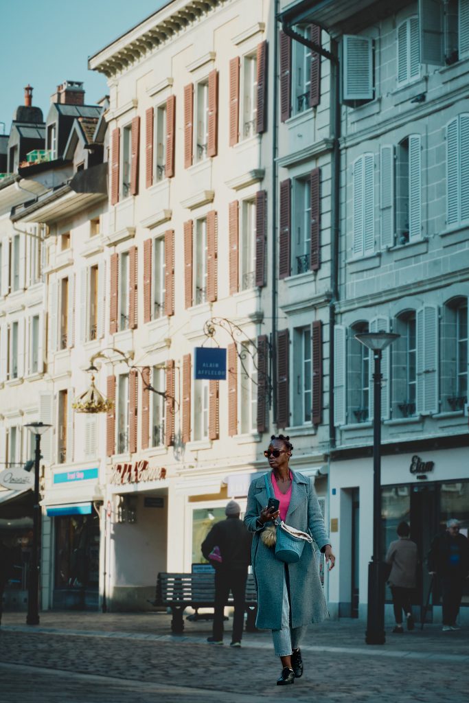 Femme élégante portant un long manteau gris et des lunettes de soleil marchant dans une rue pavée bordée de commerces et de bâtiments à l'architecture classique avec des volets.