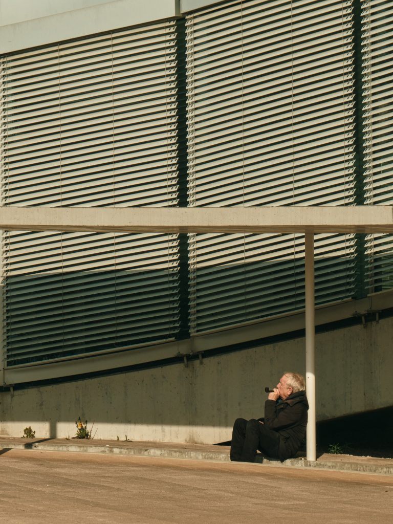 Homme assis seul au sol près d'un grand bâtiment moderne aux stores métalliques, profitant d'un rayon de soleil.