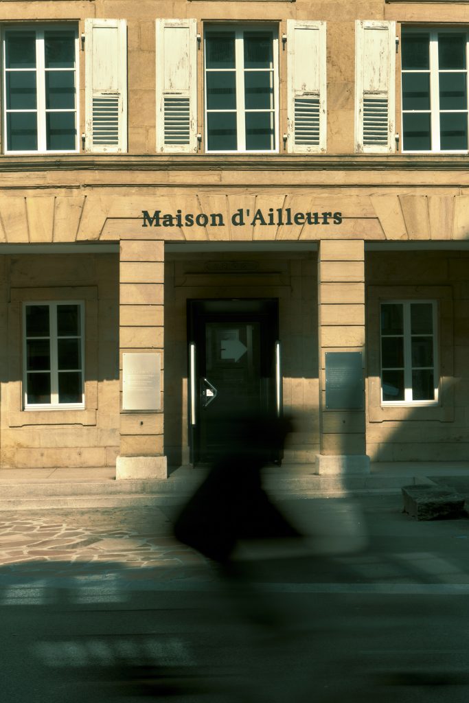 Silhouette noire et floue d'une personne passant rapidement devant l'entrée ensoleillée du musée de la Maison d'Ailleurs, un bâtiment classique en pierre de taille.