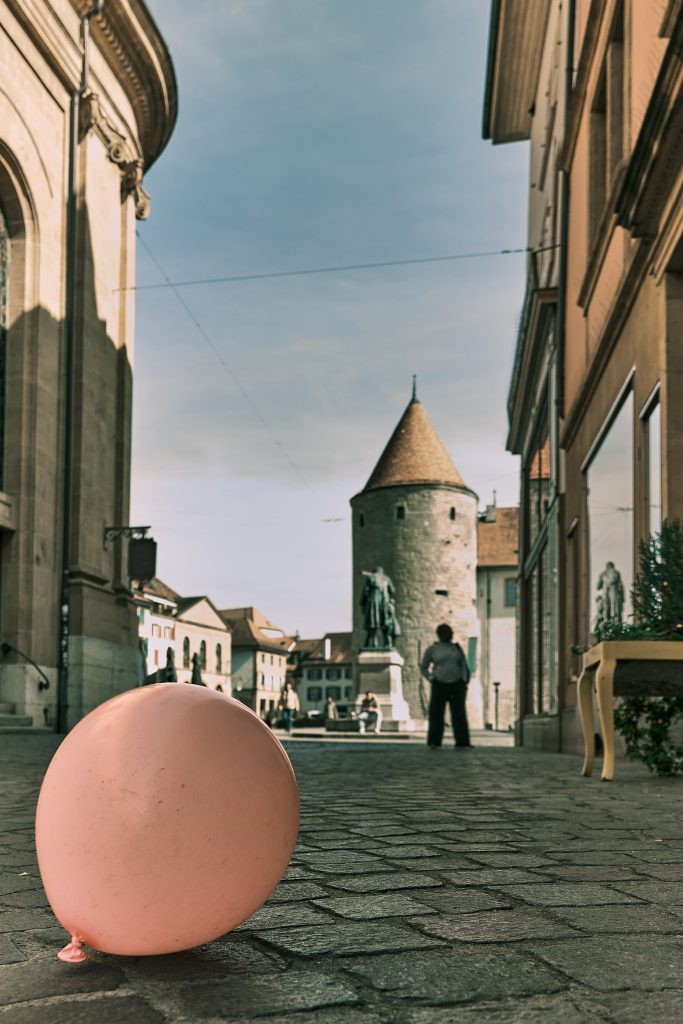 Gros plan sur un ballon rose posé sur les pavés d'une rue historique, avec une tour médiévale et des passants en arrière-plan.