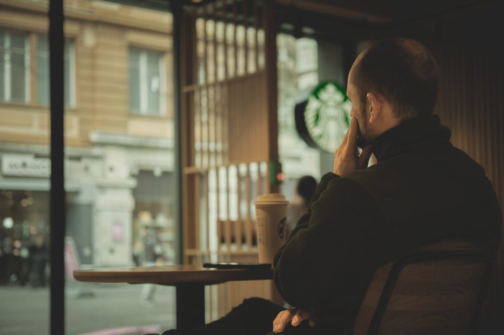 Homme assis seul à une table dans un café Starbucks, regard pensif tourné vers la fenêtre donnant sur une rue animée.