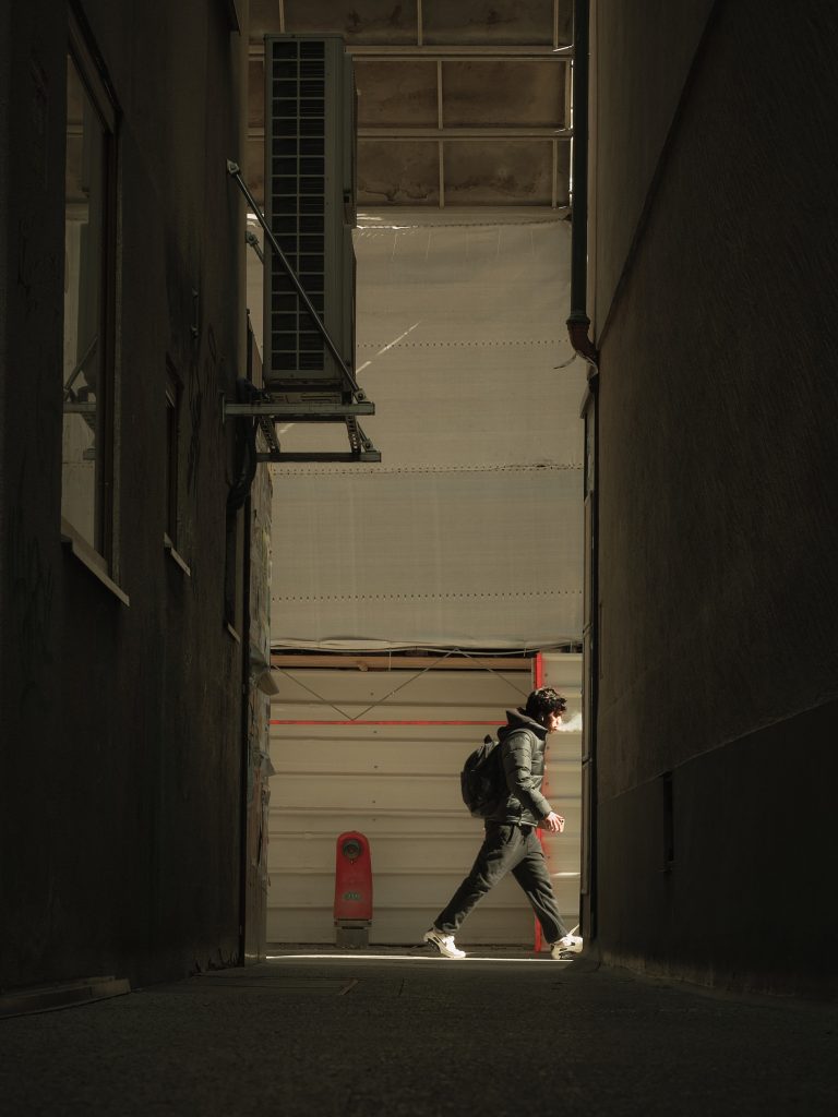 Silhouette d'une personne avec un sac à dos marchant dans une ruelle étroite et sombre, éclairée par un faisceau de lumière naturelle, exhalant de la vapeur.