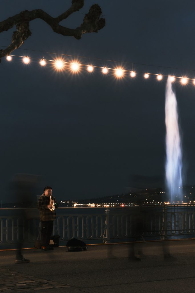 Musicien de rue jouant du saxophone de nuit au bord du lac avec le Jet d'Eau illuminé en arrière-plan.