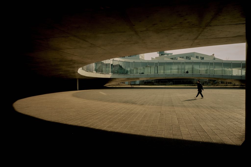 Piéton marchant sur une vaste place urbaine encadrée par une structure architecturale moderne en béton courbe.
