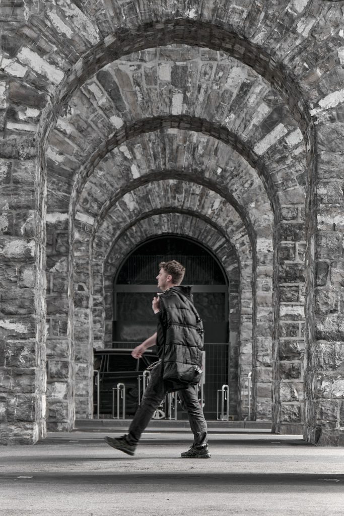 Un homme portant une veste noire marche sous une série d'arches en pierre ancienne, vue de profil.