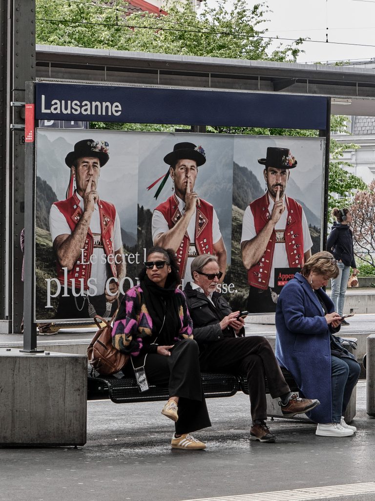 Personnes assises à un arrêt de transport en commun devant une affiche publicitaire rétro à Lausanne.