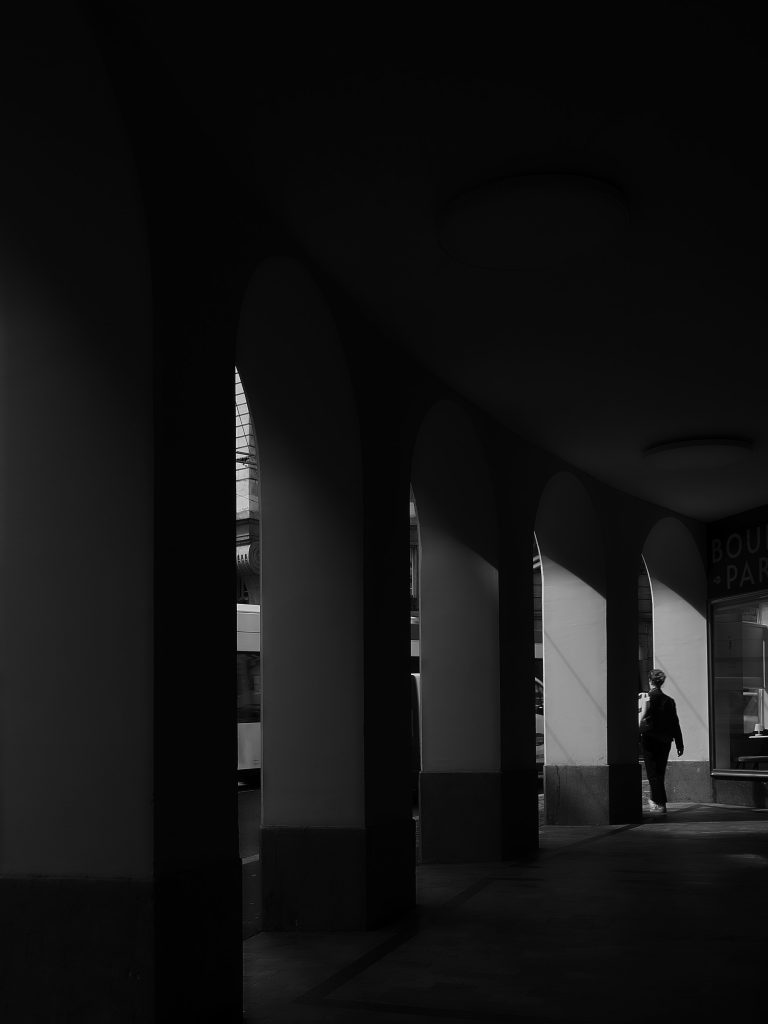 Photographie en noir et blanc d'une personne marchant sous des arcades urbaines contrastées par la lumière.