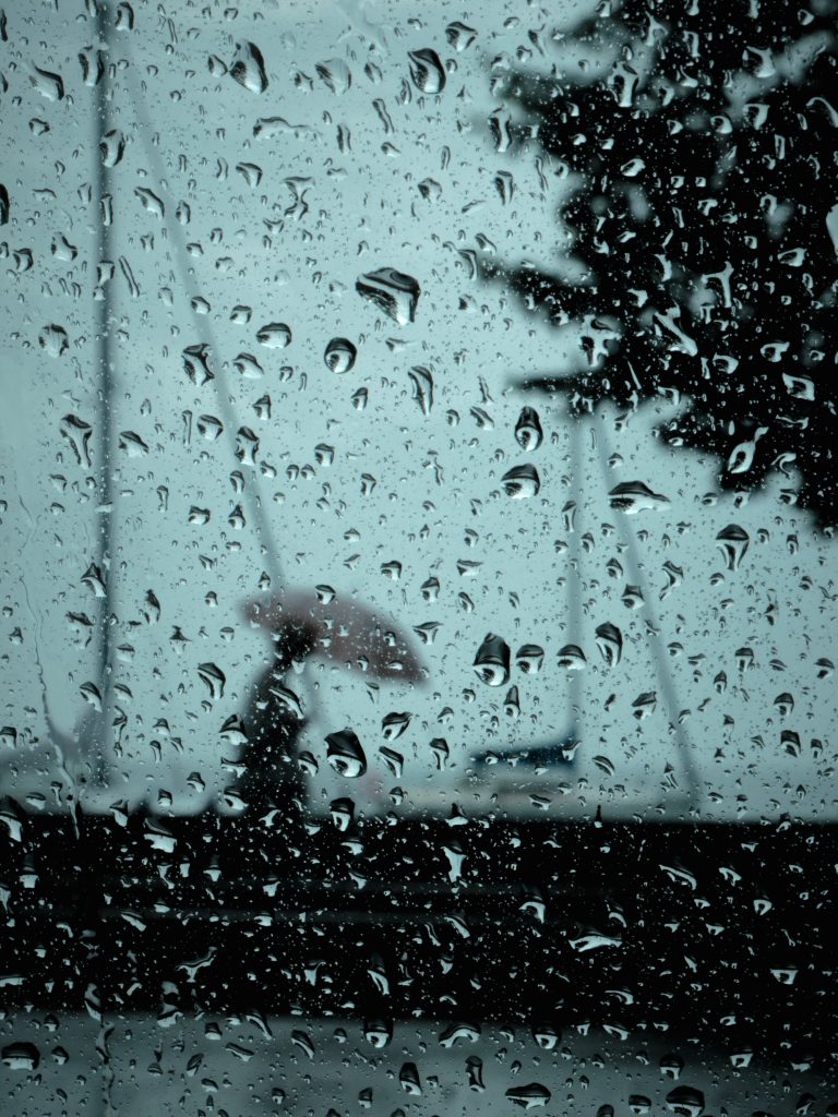 Gouttes de pluie sur une vitre avec la silhouette floue d'une personne tenant un parapluie rose en arrière-plan.