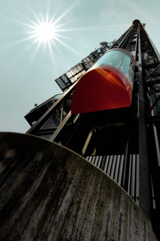Ascenseur panoramique moderne vitré avec toit rouge s'élevant le long d'une structure métallique face au soleil éclatant.