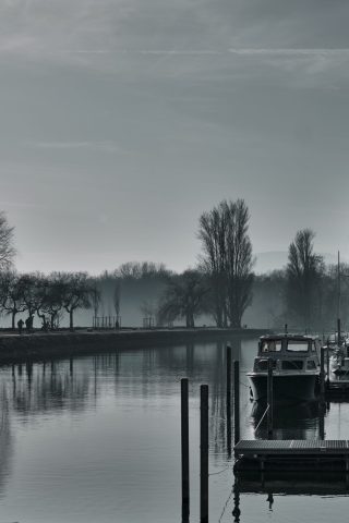 Atmosphère brumeuse sur un plan d'eau avec embarcation à quai et silhouettes d'arbres en arrière-plan.