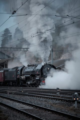 Locomotive à vapeur noire en gare dégageant d'épaisses fumées blanches sur les voies ferrées.