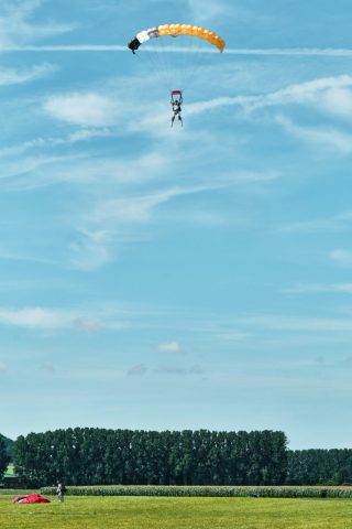 Parachutiste en plein vol sous un parachute jaune et noir descendant vers un champ verdoyant sous un ciel bleu.