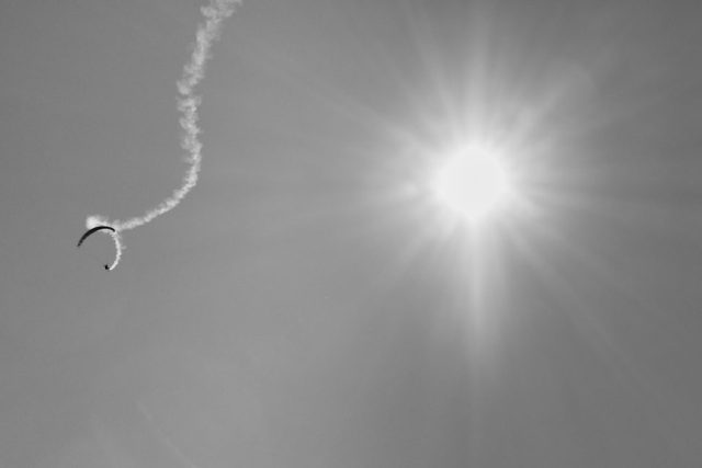 Photo en noir et blanc d'un parapentiste laissant une traînée de fumée sinueuse dans le ciel face à un soleil radieux.