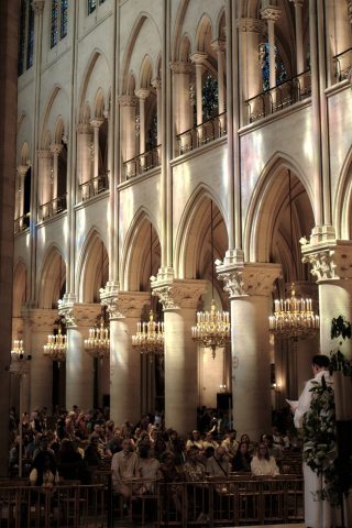 Intérieur de la cathedrale Notre Dâme à Paris avec des fidèles assis écoutant un prêtre devant de grandes colonnes gothiques et des lustres éclairés.