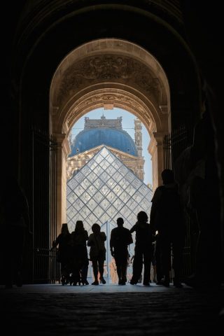 Silhouette de touristes traversant une arche de pierre avec vue sur la pyramide en verre du Musée du Louvre à Paris.