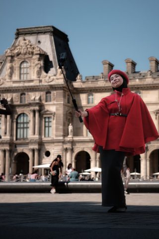 Une femme en manteau et béret rouge prend un selfie avec une perche devant la façade historique du Palais du Louvre à Paris.