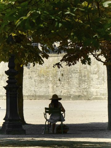 Silhouette d'une femme avec un chapeau de paille assise sur une chaise verte iconique à l'ombre des arbres d'un parc parisien.