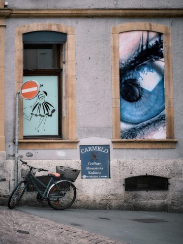 Photographie de rue d'un vélo garé devant un bâtiment ancien avec une enseigne de coiffeur et une grande affiche d'un œil bleu à la fenêtre