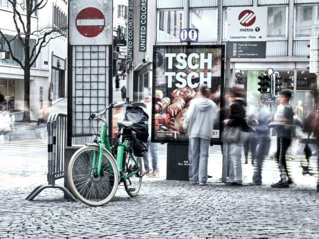 Photographie de rue avec flou de mouvement près de la station de métro Flon à Lausanne, montrant des piétons, un vélo vert et un panneau publicitaire