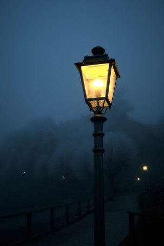 Réverbère vintage allumé diffusant une lumière chaude dans un parc de nuit sous une atmosphère brumeuse et mystérieuse.