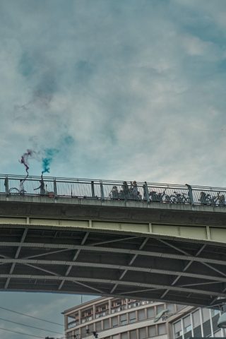 Personnes rassemblées sur un grand pont urbain allumant des fumigènes de couleur bleue et violette sous un ciel nuageux.