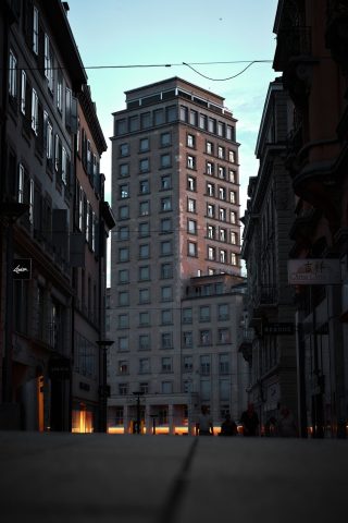 Grande tour en pierre éclairée au crépuscule au bout d'une rue piétonne encadrée par des bâtiments urbains sombres.
