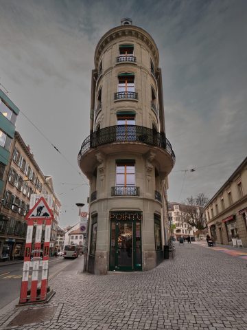 Bâtiment d'angle arrondi classique abritant le café Le Pointu, au croisement de deux rues pavées sous un ciel nuageux.